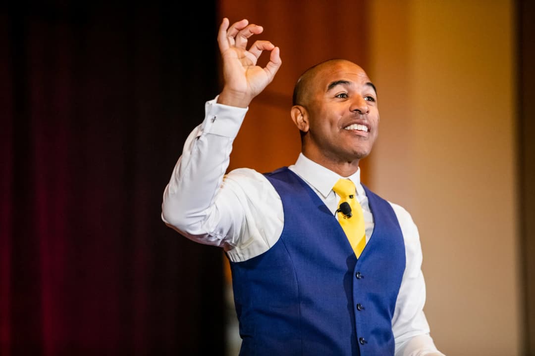 A speaker in a blue vest and white shirt is gesturing with one hand raised in an 'okay' sign while smiling and engaging with the audience. The background features warm colors suggesting a conference or event setting.