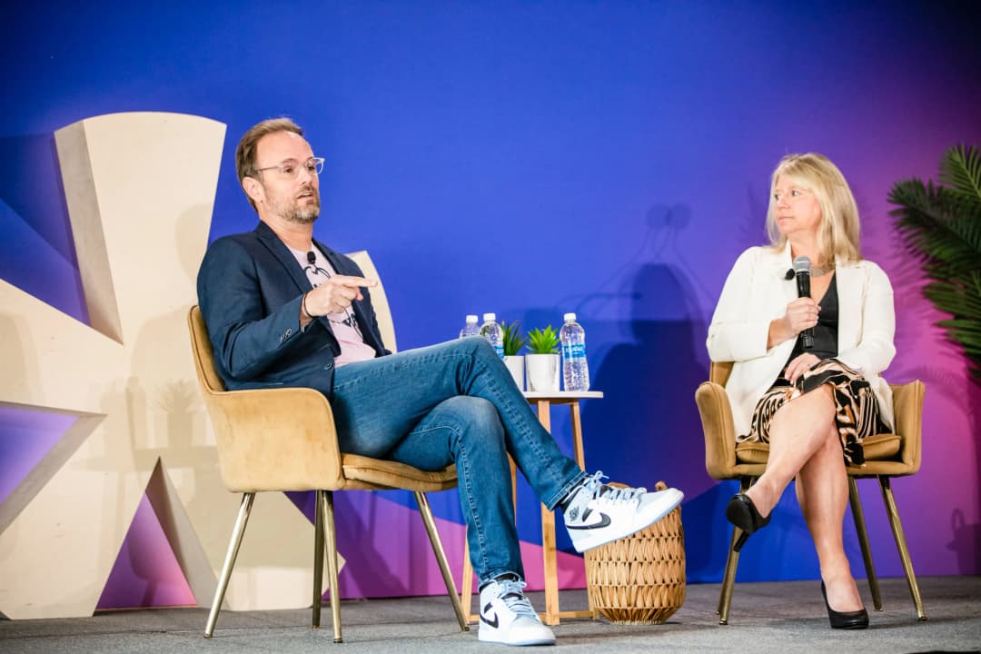 A man and a woman are seated on a stage, engaged in a discussion at an event. The man is wearing a navy blazer over a light t-shirt, paired with jeans and athletic shoes. He is gesturing while speaking. The woman, dressed in a black top and a patterned skirt, is holding a microphone and looking attentively at the man. In the background, there are two bottles of water and a small potted plant on a table between them, set against a gradient blue and purple backdrop. A large decorative element is visible to the left of the image.