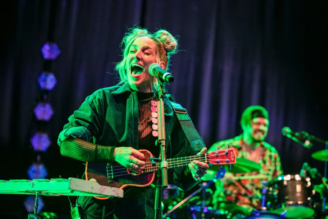 A performer passionately singing into a microphone while playing a small guitar on stage, with vibrant green lighting illuminating their face. Behind them, a drummer can be seen in the background, contributing to the lively atmosphere of the performance. The backdrop features dark drapery, highlighting the colorful stage lighting.