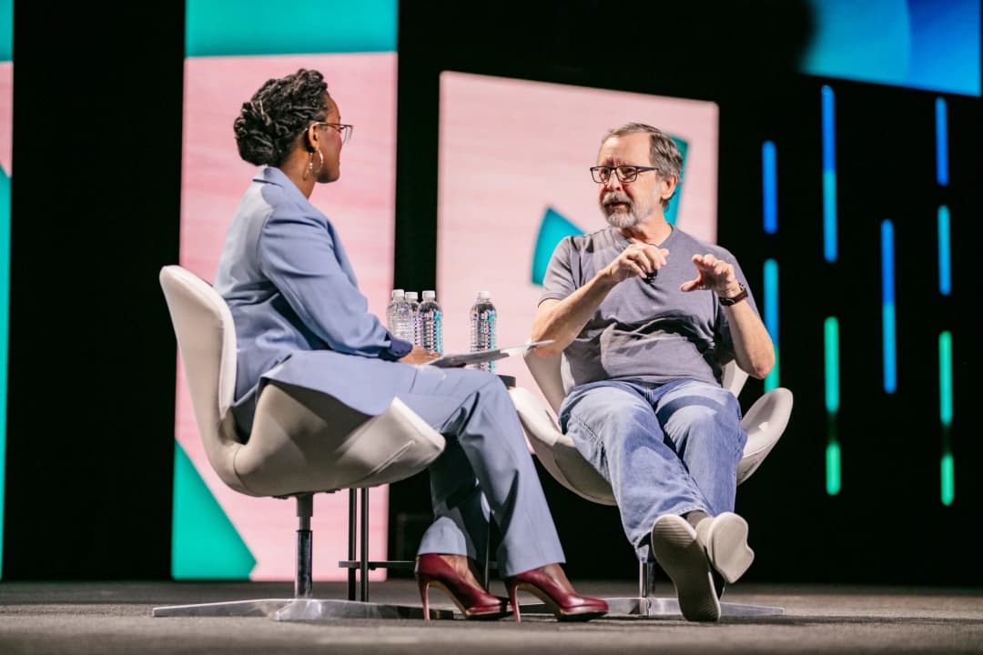 A woman in a blue suit and glasses and a man in a gray shirt are engaged in a discussion on stage at an event. The woman is seated in a modern chair, while the man, speaking animatedly, gestures with his hands. On the table between them are bottles of water. The backdrop features colorful geometric designs, creating a lively atmosphere for their conversation.