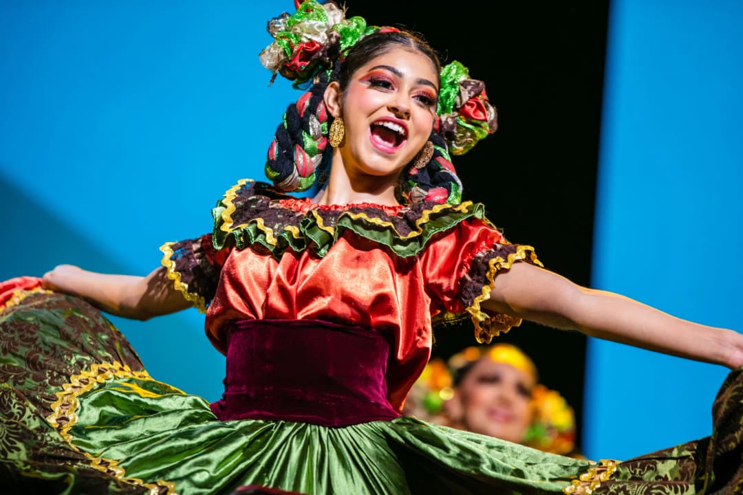 A young woman is performing on stage, dressed in a vibrant traditional costume featuring bright colors like red and green, with frilly edges and floral decorations. She has long, braided hair adorned with colorful accessories, and she is joyfully smiling while showcasing her dance. In the background, a second performer can be seen, adding to the lively atmosphere of the performance. The backdrop is a bright blue, enhancing the colors of the dancers' attire.