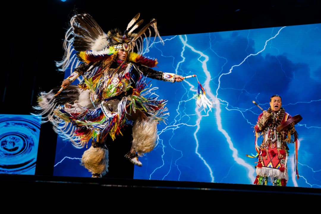 A performer dressed in colorful traditional attire dances energetically on stage, showcasing vibrant feathers and beaded decorations. In the background, a dynamic display features lightning effects against a blue backdrop. Another individual, also in traditional dress, plays a drum while singing, contributing to the lively atmosphere of the performance.