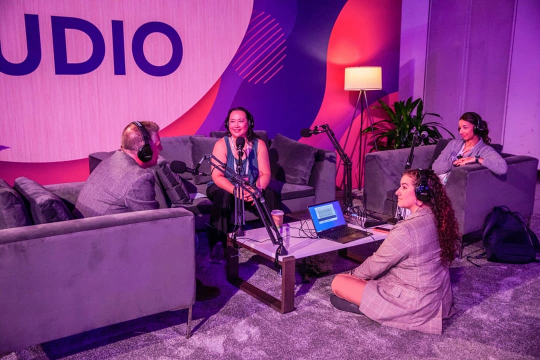 A group of four people engaged in discussion in a podcast studio setting. Two individuals, one male and one female, sit on gray couches with microphones in front of them, while another woman listens attentively. A laptop and a coffee cup are placed on a table between them. The background features vibrant colors with the word 'STUDIO' prominently displayed. The atmosphere is casual and collaborative.