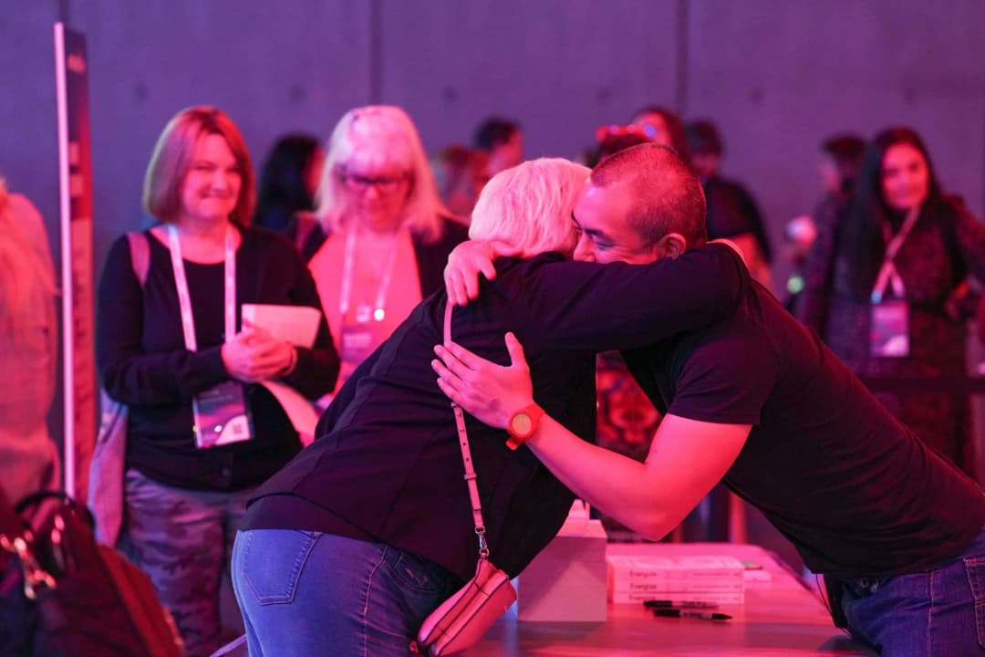 A warmly lit gathering featuring a man embracing a woman, both smiling as they share a heartfelt moment. In the background, a group of attendees, including three women holding items, watch the interaction. The setting conveys a sense of connection and camaraderie among participants at an event.