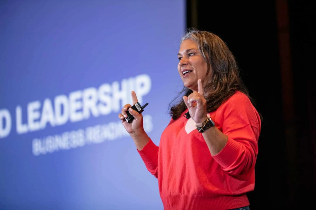A presenter in a red sweater gestures while speaking on stage. The backdrop displays the text 'LEADERSHIP' in large letters. The presenter holds a remote clicker and appears engaged in their presentation, addressing an audience not visible in the image.