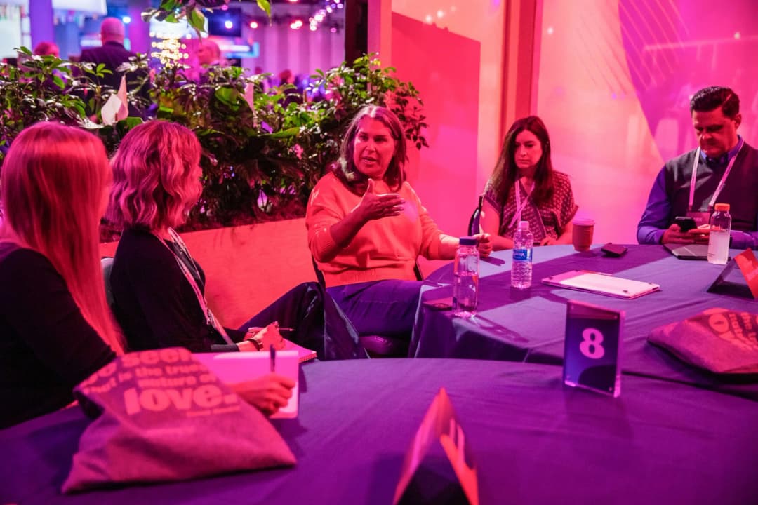 A small group of participants engaged in a discussion around a table during a conference. The scene features a woman in an orange sweater animatedly speaking, while other attendees listen attentively, with some taking notes. The setting is colorful, with purple lighting and foliage in the background, and a table covered with a blue cloth displaying drinks and a number marker (8).