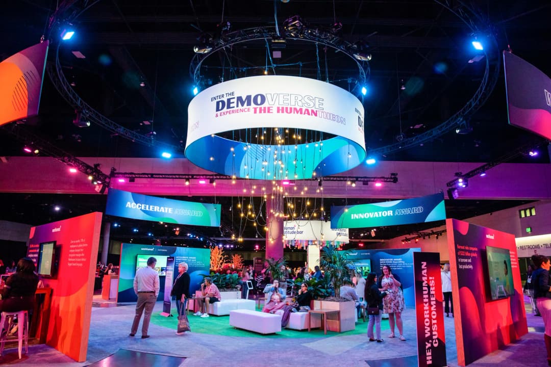 A vibrant exhibit space at the Workhuman Live conference showcases the Demoverse, with colorful signage promoting the 'Accelerate Award' and 'Innovator Award.' Attendees engage in conversation among lush greenery and modern seating arrangements, while bright lights enhance the atmosphere. The decor features various banners with dynamic colors and promotional content, creating an inviting and energetic environment for networking and exploration.