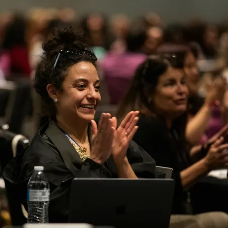 A woman with curly hair smiles and claps enthusiastically while seated in a crowded conference or event. In the foreground, a bottle of water and a laptop are visible, suggesting an engaged audience. A woman sitting beside her also appears interested in the presentation. The atmosphere conveys positivity and excitement.