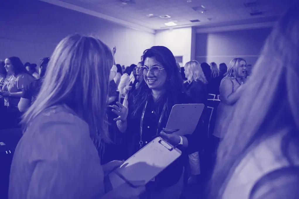 A group of individuals engaged in conversation at an event, with two women at the forefront interacting closely. One woman appears to be smiling and holding a clipboard while the other is listening intently. In the background, other attendees can be seen mingling, creating a lively atmosphere.
