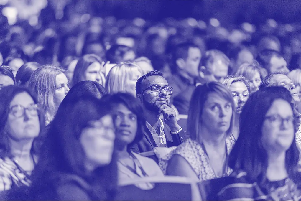 A diverse audience is seated and attentively watching a presentation, with one man in the foreground, resting his chin on his hand, showcasing a thoughtful expression. The image is presented in shades of blue, giving it a unique visual effect.