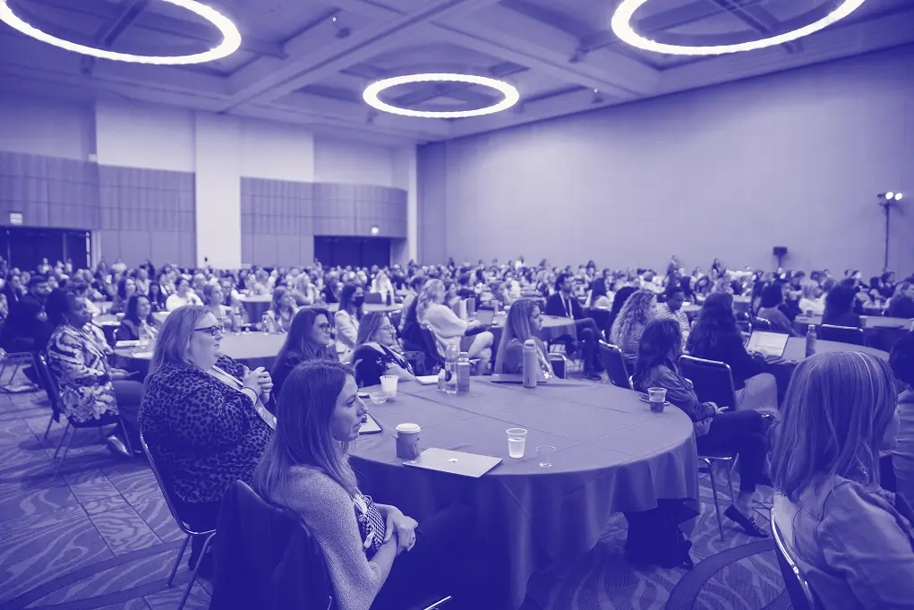 A large audience attends a conference in a spacious room, seated at round tables. The atmosphere is engaged, with many individuals looking towards the front of the room. Bright circular lights hang from the ceiling, highlighting the attendees, who are predominantly women. Some people are taking notes or using devices on the tables in front of them.