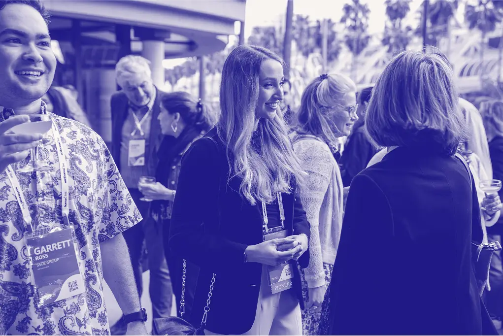 A group of attendees engaging in conversation at an outdoor event. In the foreground, a man in a patterned shirt smiles while holding a drink. Nearby, a woman with long hair wearing a dark outfit chats with another attendee. The background shows a mix of people, some with drinks, mingling in a lively atmosphere, surrounded by palm trees and a blurred architectural feature. Name tags can be seen hanging from some guests' necks.
