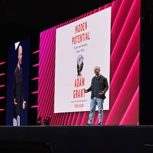 A speaker, Adam Grant, stands on stage presenting with a vibrant backdrop featuring the title of his book, "Hidden Potential." The screen highlights his name and the book's cover, which includes a visual element related to the theme. The stage is set with dramatic lighting, enhancing the presentation atmosphere.