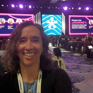 A smiling woman takes a selfie in an event space, with large screens in the background displaying colorful graphics and text that reads "Ready on the JOURNEY." The venue features rows of seats filled with attendees, illuminated by purple and blue lights.