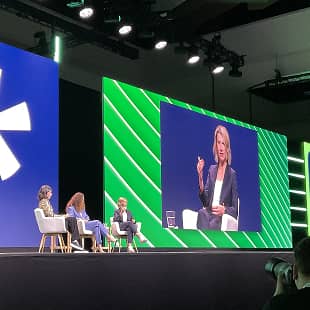 A panel discussion featuring three speakers on stage at a Workhuman event. One speaker is engaged in conversation, gesturing while the other two listen intently. The background consists of large screens displaying a stylized logo and graphic design elements. The setting is well-lit with an audience visible in the foreground, indicating an interactive event atmosphere.