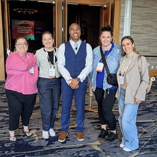A group of five attendees posing together for a photo at a conference. They are smiling and standing in front of an entrance with event-related displays and banners visible in the background. The attendees are wearing a mix of professional and casual attire, with name badges attached to their clothing. The ambiance suggests a networking or social event.