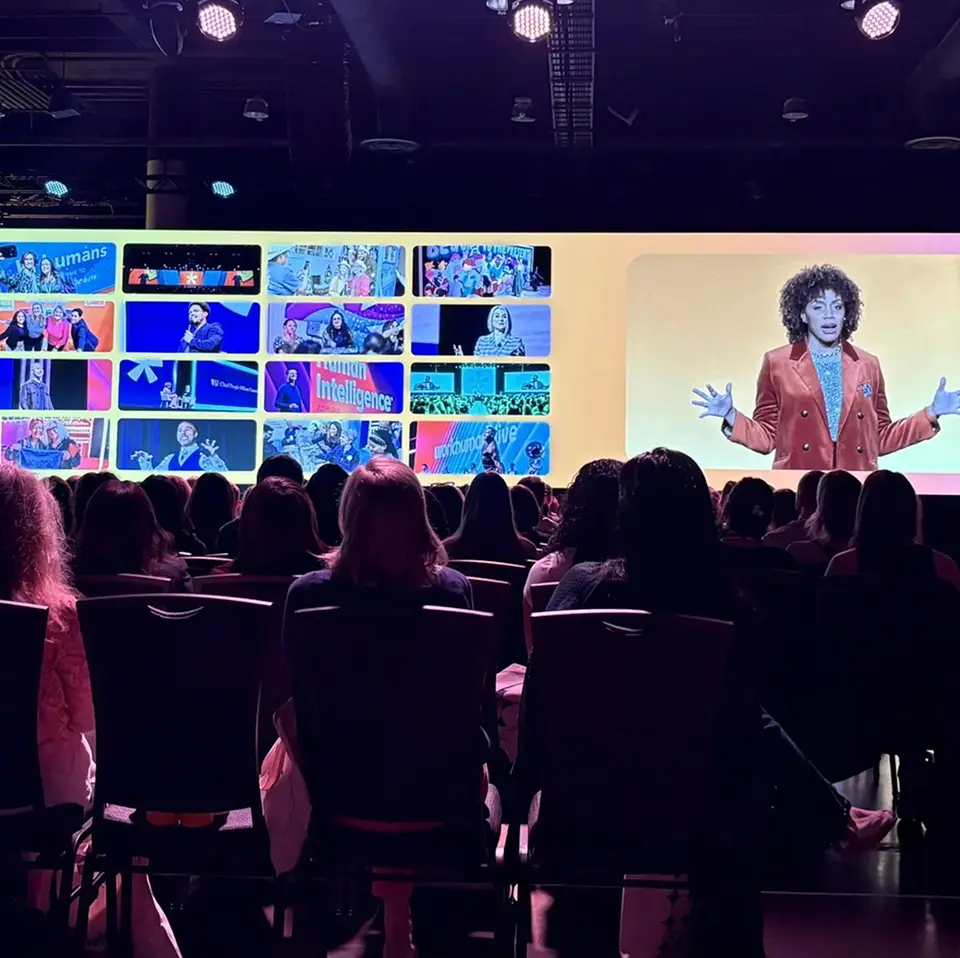 A speaker in a vibrant orange blazer gestures while addressing an audience from a large screen displaying various images related to the Workhuman event. The audience, mostly women, is visible in the foreground, watching attentively. The screen features a collage of moments from the event, including different speakers and activities, capturing the engaging atmosphere of the conference.