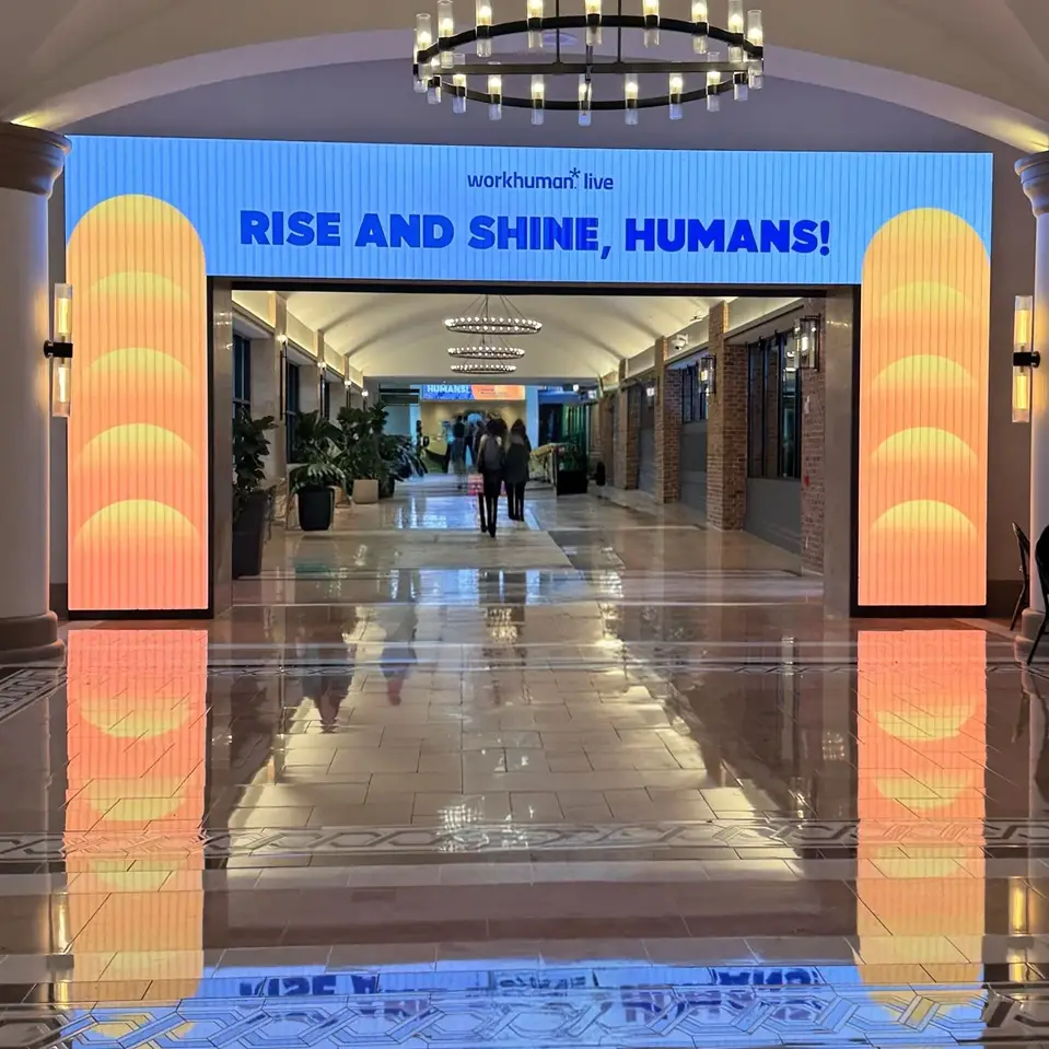 A brightly lit hallway entrance for the Workhuman Live event features a large digital display with the text "RISE AND SHINE, HUMANS!" at the top. The entrance is framed by colorful, glowing panels, while attendees walk down the corridor, which has polished floors reflecting the lights and surroundings. Lush plants are placed along the sides, contributing to the inviting atmosphere of the event space.