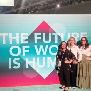 A group of three women posing in front of a large mural that reads "THE FUTURE OF WORK IS HUMAN." The backdrop features a colorful gradient with diamond shapes, emphasizing a modern and vibrant atmosphere. The women are smiling and dressed in professional attire, showcasing an engaging setting.