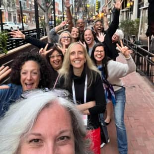 A group of people are having fun outdoors as they pose for a selfie. There are several individuals in the background, displaying cheerful expressions and waving their hands. The scene is lively, with many smiles and gestures of excitement, creating a sense of camaraderie among the group. The setting appears to be a city street, suggesting a social or professional gathering.