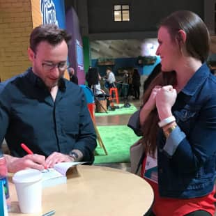A man is seated at a table, writing in a notebook with a red pen while a woman, standing beside him, appears excited or happy, clenching her hands in front of her. The background features colorful elements typical of a conference or event setting, with attendees visible in the distance.