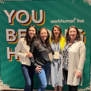 A group of four women smiling together in front of a colorful backdrop that reads "YOU belong HERE" alongside the Workhuman Live logo. Each woman is dressed casually, with two wearing jeans and two in dresses, showcasing a friendly and welcoming atmosphere.