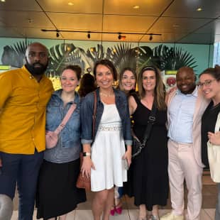 A group of six people posing together for a photo in a brightly lit interior, with a decorative background featuring plants and modern design elements. The group consists of three women and three men, each wearing stylish and casual outfits, smiling at the camera.