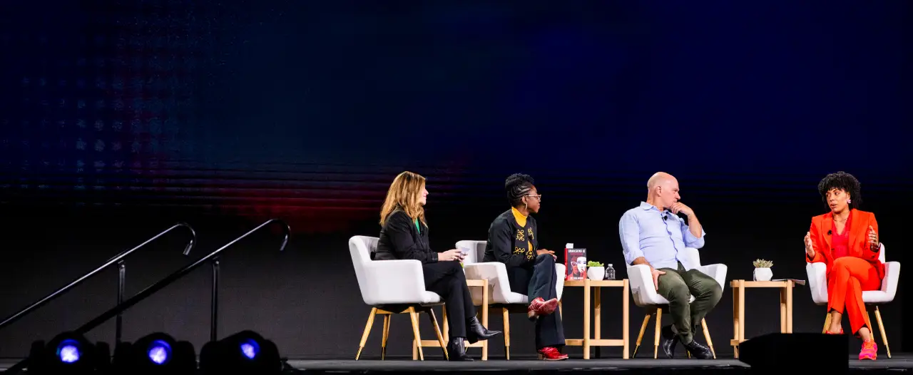 A panel of four speakers discussing a topic at a Workhuman event, with a large backdrop displaying the Workhuman logo. The panel includes a person in an orange outfit, another in a black top, and a third in a blue shirt, while the fourth speaker sits in a black outfit. Each speaker has a microphone and there are small tables with items in front of them. The audience is visible in the foreground, and the stage is well-lit, emphasizing the speakers.