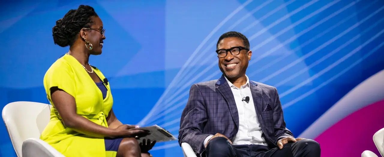 A woman in a bright yellow dress and glasses smiles while seated next to a man in a dark suit and glasses, who appears to be laughing. They are engaged in conversation on a stage with a blue and pink abstract background. The woman holds a small item in her hand, and they both appear friendly and approachable.