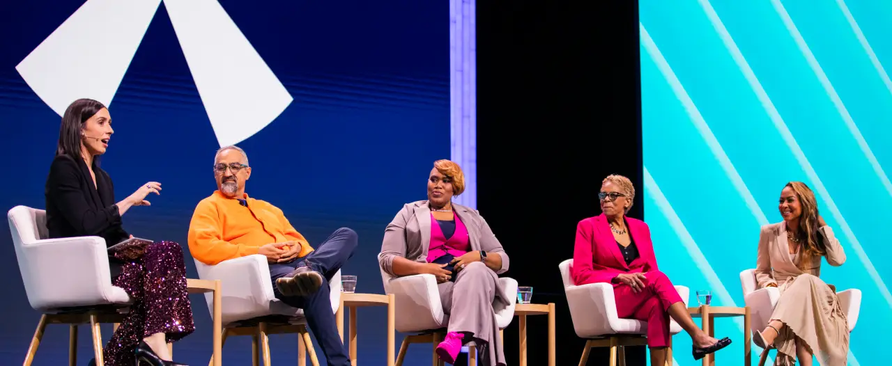 A panel discussion featuring five speakers on stage at a Workhuman event. They are seated in modern chairs, engaged in conversation, with a colorful backdrop of abstract shapes. The speaker on the left has long dark hair and is wearing a black top and sparkly pants, while the others are dressed in a variety of styles, with one wearing an orange sweater, another in a gray outfit with pink accents, and two in professional dresses. The scene captures a moment of dialogue at the event.