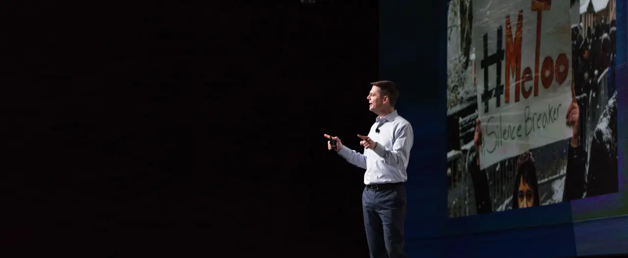 A speaker stands on stage with a spotlight, gesturing while presenting. Behind him, there is a large screen showing a crowd holding a sign with the text "#MeToo" and "Silence Breaker." The speaker appears engaged and is addressing the audience, highlighting the significance of the message displayed.