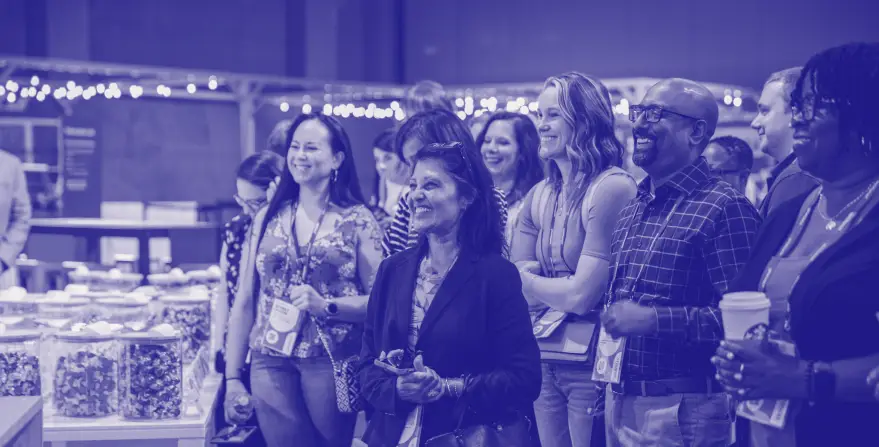 A group of diverse attendees smiling and engaging at an event, gathered in front of display tables filled with jars. The setting is lively, with warm lighting and a sense of camaraderie among the participants.