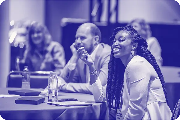 A woman with long, curly hair is smiling while engaged in a conversation at a table. She is wearing a white blazer and holding a pen, expressing interest in the discussion. In the background, a man sits with his chin resting on his hand, showing attentiveness, while another woman can be seen behind them, also engaged in the event. There are water bottles and notebooks on the table, indicating a collaborative setting.