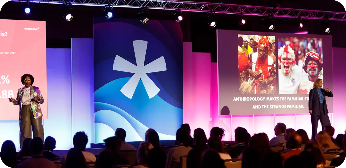 A speaker presenting at a Workhuman event stands on stage wearing a colorful blazer, gesturing towards a screen that shows an image of people in cultural attire, as well as text stating, "Anthropology makes the familiar strange and the strange familiar." In the background, an audience is seated, engaged in the presentation. The stage features bright pink and blue lighting along with the Workhuman logo.