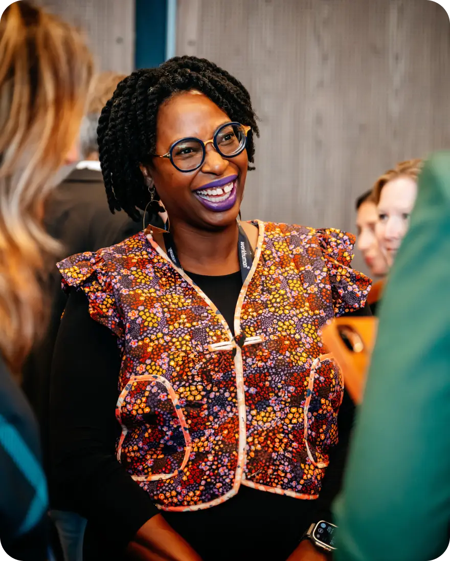 A smiling woman wearing a colorful patterned top stands engaged in conversation, with other attendees in the background. She has curly hair and large glasses, and her joyful expression conveys a sense of connection and excitement at the event. The setting appears to be a social gathering, with warm lighting and modern decor.