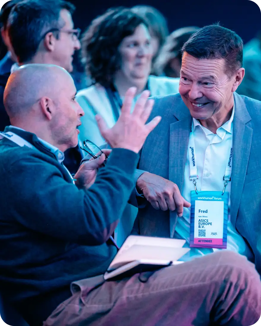 Two men are engaged in a lively conversation at a Workhuman event. One man, wearing a gray sweater, gestures with his hands, possibly emphasizing a point, while the other man, dressed in a light gray suit, smiles in response. In the background, several attendees can be seen, indicating a busy event atmosphere. A name tag hanging from the suit identifies one of the men as "Fred van Wees" from ASICS EUROPE B.V.