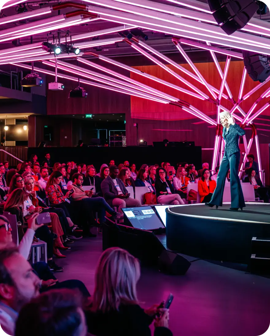 A speaker stands on a stage illuminated by vibrant pink and white lighting, addressing an audience that is seated in a large venue. The audience members appear engaged, with some holding their smartphones. The stage is circular with a display showing a timer, and the overall atmosphere suggests a dynamic and interactive event.
