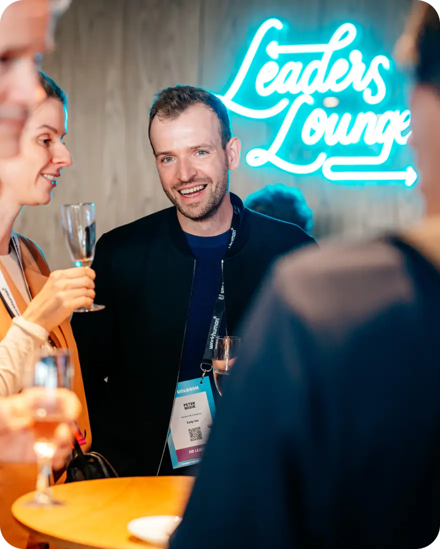 A smiling man stands in a social setting, holding a drink, while chatting with a woman beside him who also has a drink. He wears a black jacket and has a name tag visible that reads "PETER Coty." Behind them is a neon sign that says "Leaders Lounge." The scene conveys a relaxed atmosphere of networking and conversation at an event.
