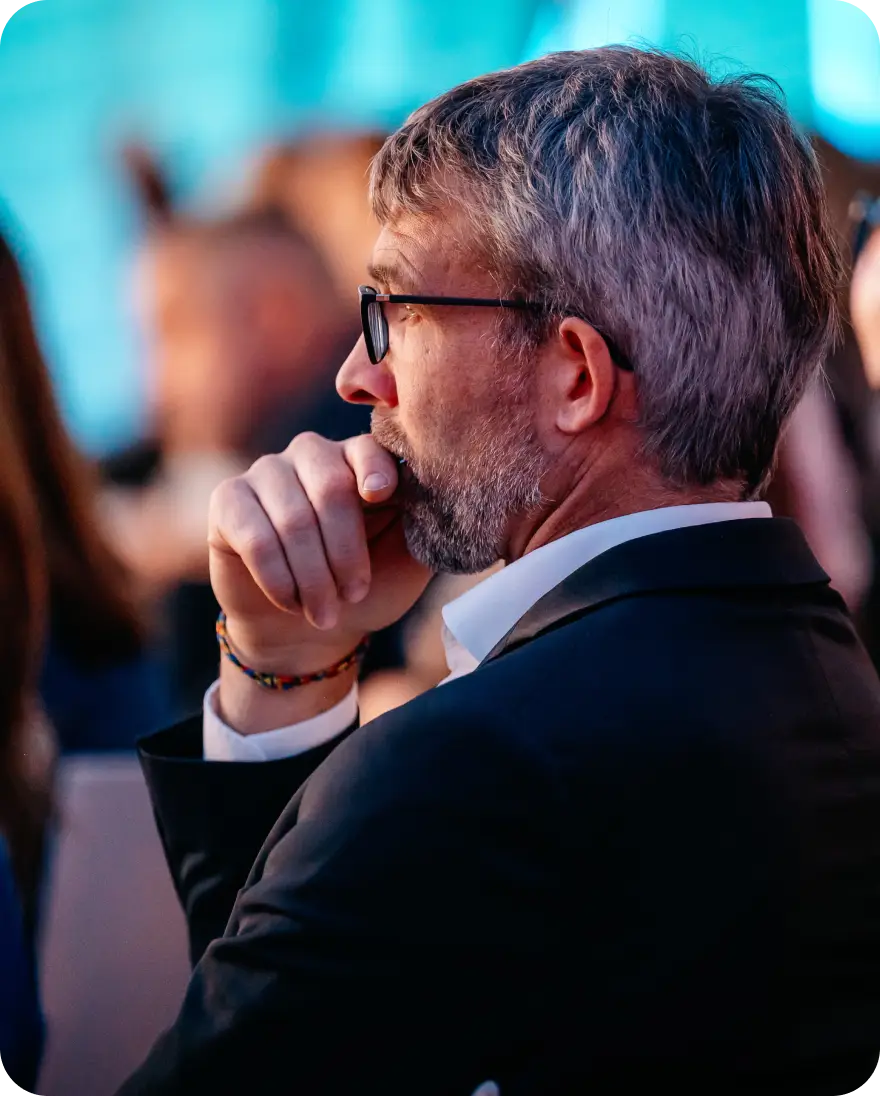 A thoughtful man with a beard and glasses rests his chin on his hand, appearing engaged while listening to a presentation or discussion. He is dressed in a suit and sits in a blurred audience environment, with soft blue lighting in the background conveying an event setting.