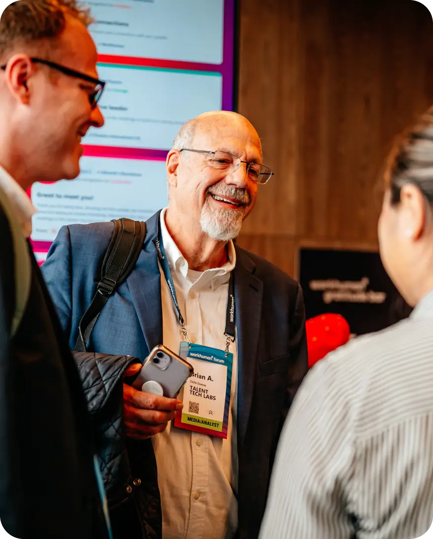A man with glasses and a beard smiles as he engages in conversation with two other individuals at a Workhuman event. He is dressed in a suit and holds a smartphone in one hand while wearing a name badge that reads "Brian A. Delle Donne, Talent Labs, Media/Analyst." In the background, colorful screens display messages related to the event, adding to the lively atmosphere.