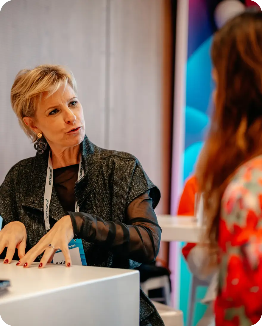 A woman with short blonde hair is engaged in conversation at a table, gesturing animatedly with her hands. She is wearing a black top with a gray overlay and has a lanyard displaying the word "workhuman." The background features colorful abstract designs, and another person is partially visible to the right, adding to the interaction's context. The setting appears to be an event or conference.