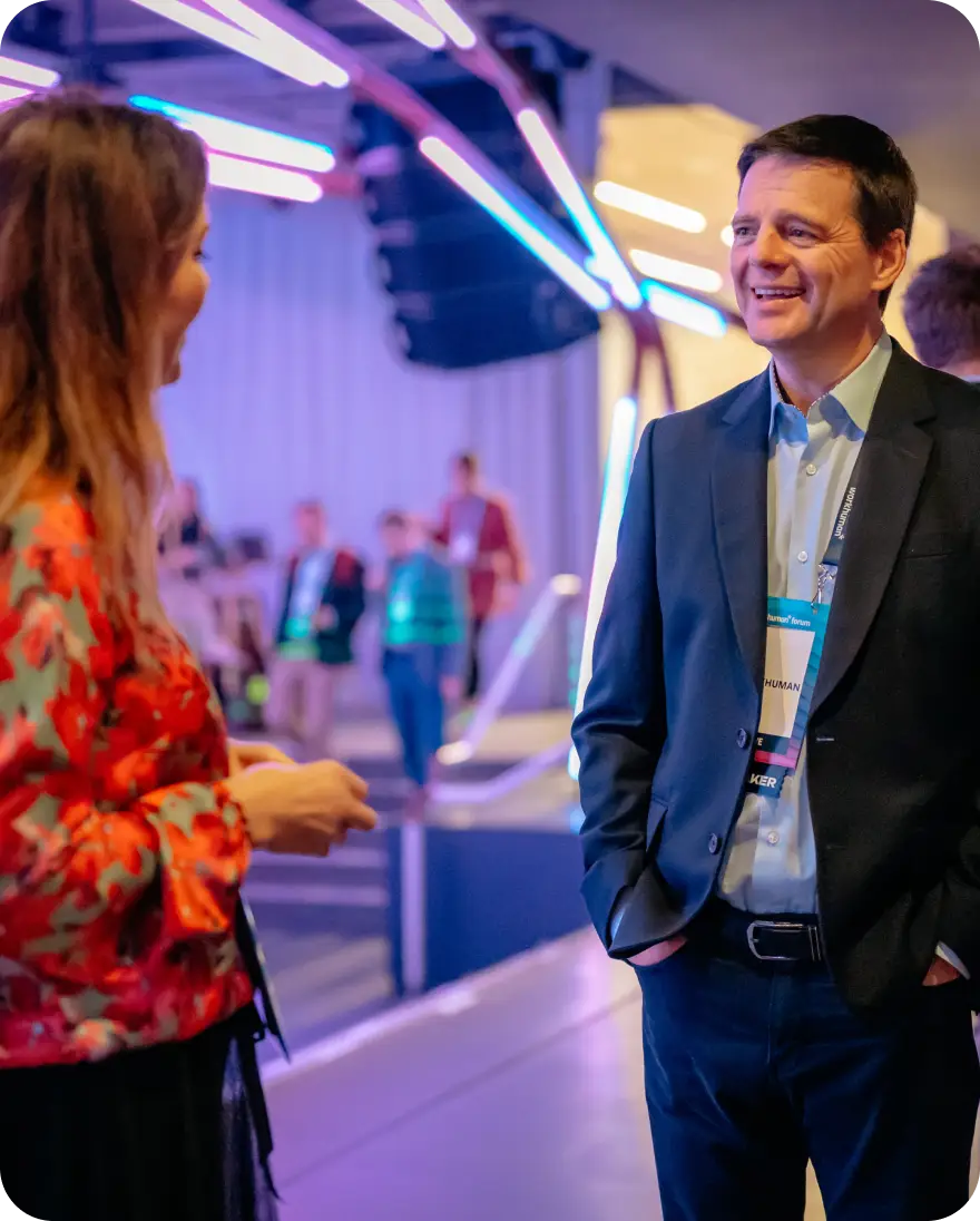 A man in a suit smiles as he chats with a woman wearing a floral blouse. They are engaged in conversation at a lively event, with colorful lighting and other attendees in the background. The atmosphere appears to be energetic and professional, likely part of a conference or gathering. The man's name tag indicates that he is associated with Workhuman.