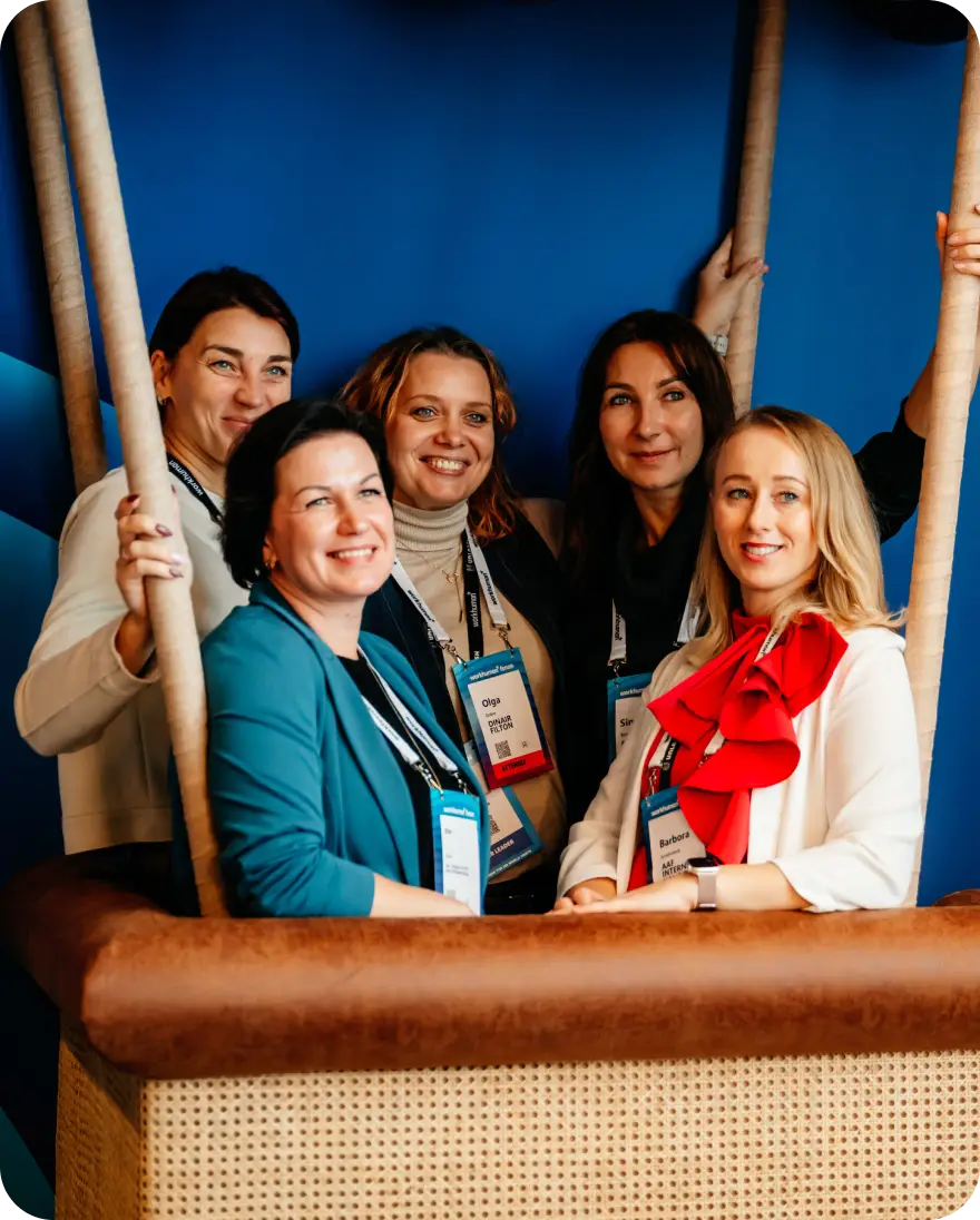 Five women pose together in a photo booth styled as a hot air balloon. They are smiling and wearing name badges. The background features a blue design, adding to the cheerful atmosphere of the image.