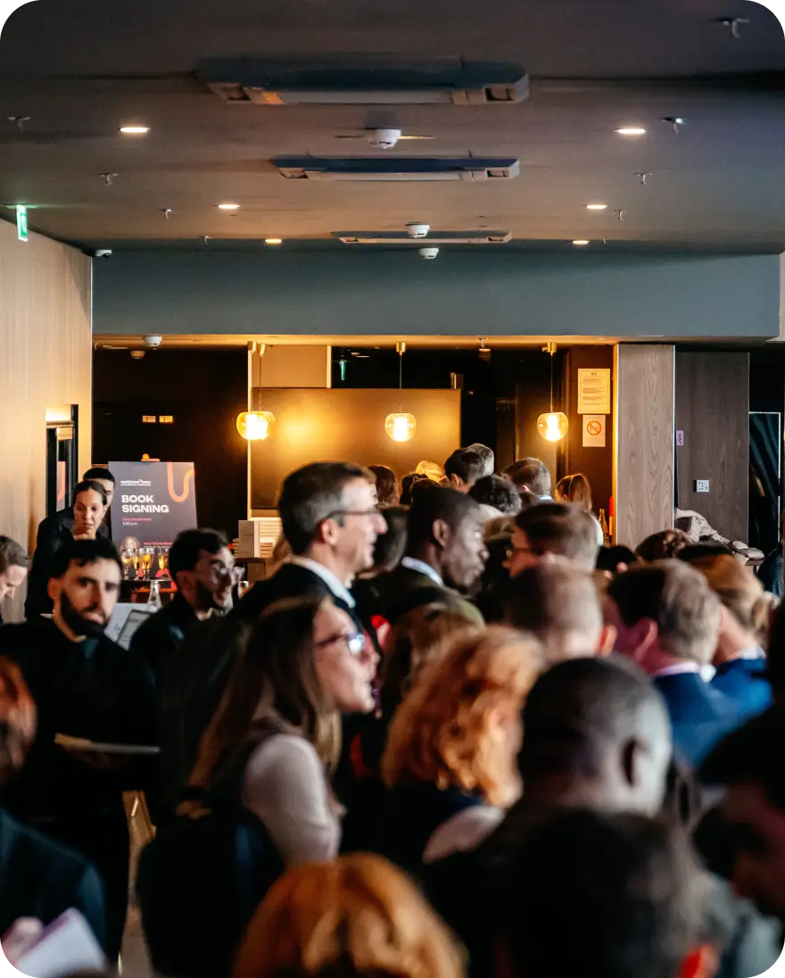 A crowded event space featuring a diverse group of attendees mingling and interacting. In the background, a sign indicates a "BOOK SIGNING" event. The space is well-lit with modern decor, including soft lighting and a stylish ambiance. Attendees are dressed in professional attire, suggesting a formal gathering such as a conference or networking event.