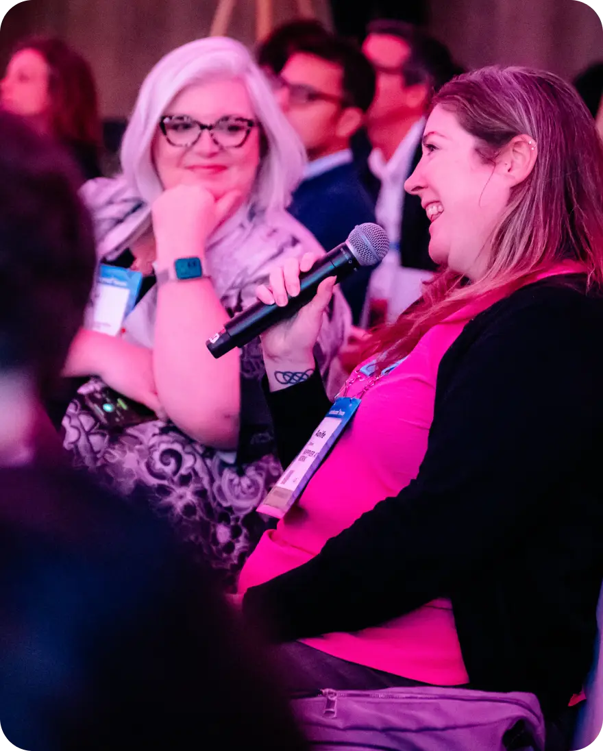 A woman with long hair, wearing a pink top, is speaking into a microphone during a discussion while another woman with glasses and white hair listens attentively, resting her chin on her hand. The background features a blurred audience amid colorful lighting. Both women are wearing name tags.