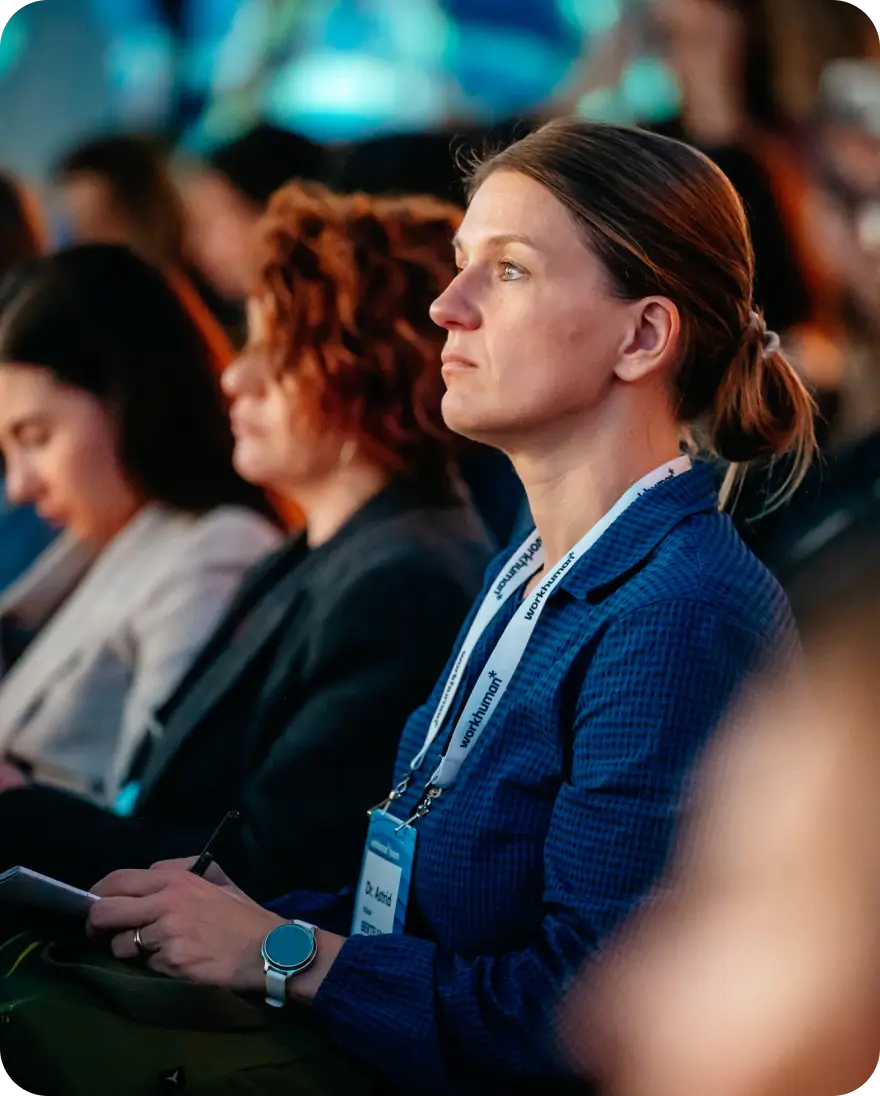 A focused woman with a ponytail sits in the audience, holding a notepad and pen, listening intently during a conference or event. She wears a blue jacket and a name badge with the text "Dr. Ivorid" prominently displayed. In the background, several other attendees are engaged in the presentation, although they are not clearly visible. The environment suggests a professional setting, likely related to Workhuman events.