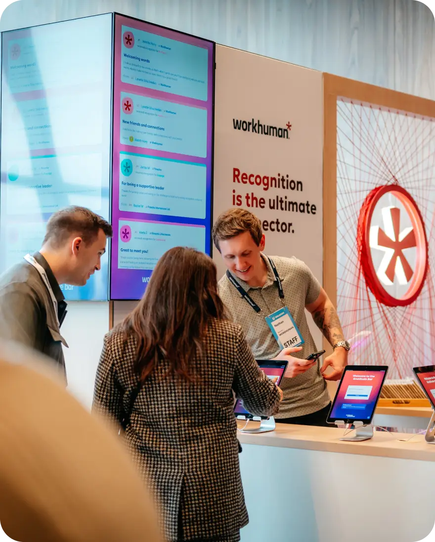 A group of people interact at a Workhuman event, looking at digital displays with recognition messages. One display shows welcoming words such as "New friends and connections" and "For being a supportive leader." A staff member with a name tag assists attendees at the information booth. The background features Workhuman branding and a decorative element.