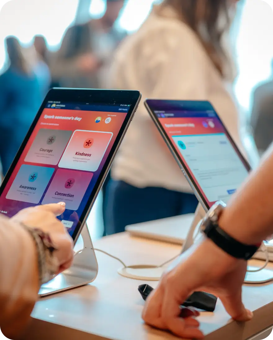 A close-up view of two tablets on a table, showcasing an interface from Workhuman. One tablet displays options for recognizing others with buttons labeled "Courage," "Kindness," "Awareness," and "Connection." A hand is poised over the screen, indicating interaction. In the background, people are engaged in conversation, creating a lively atmosphere.