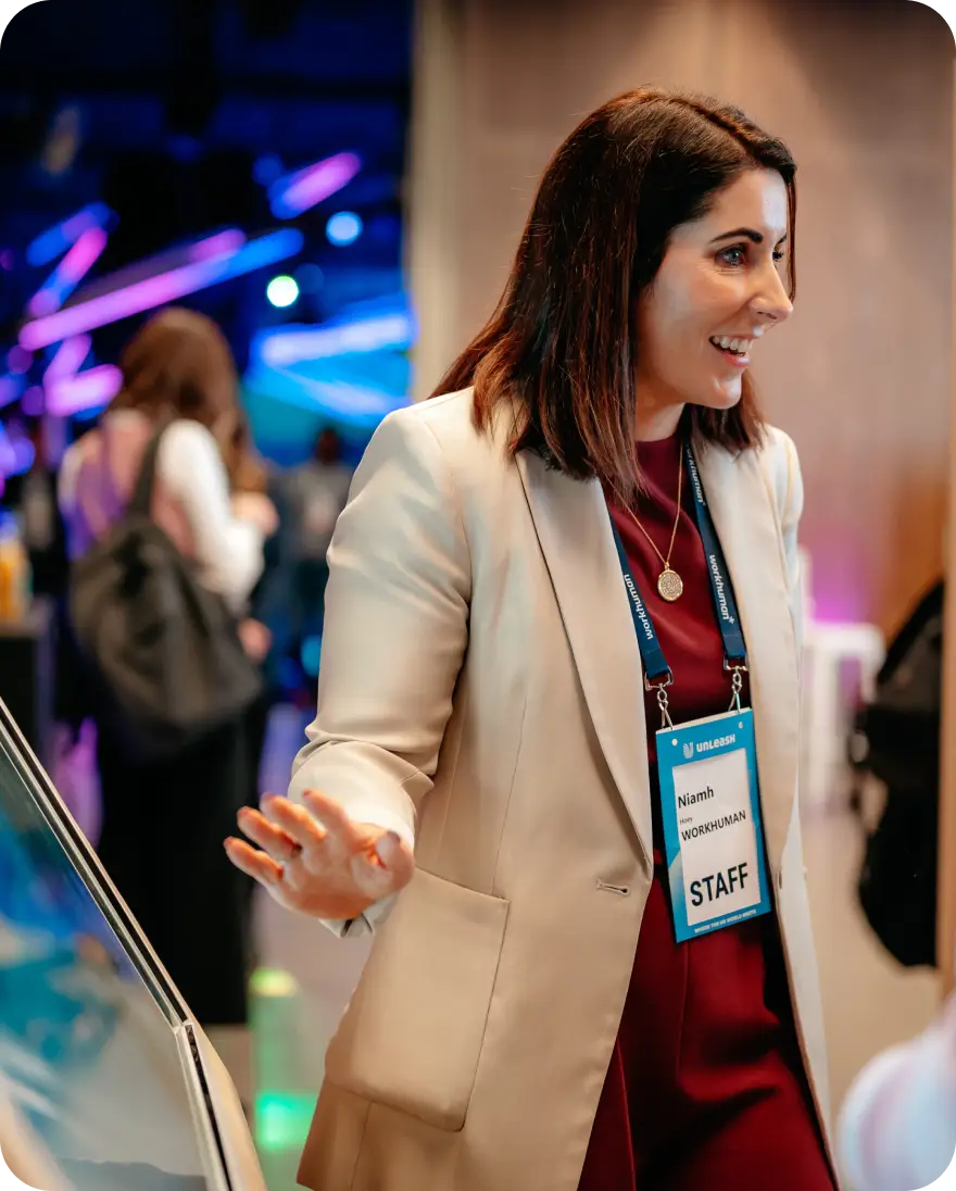 A woman with medium-length dark hair is smiling and interacting with attendees at an event, wearing a light-colored blazer over a maroon top. She has a name tag that reads "Niamh - WORKHUMAN STAFF." The background features colorful lighting and blurred figures of people, creating a lively atmosphere.