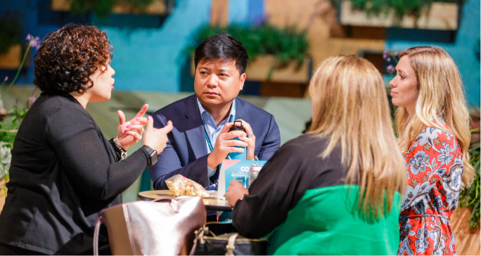 A group of four professionals engaged in conversation at a round table, with one woman animatedly gesturing while speaking. The man in the center appears to be listening attentively, holding a phone, while two other women are also involved in the discussion. The background features a colorful, plant-adorned setting, creating a vibrant atmosphere.
